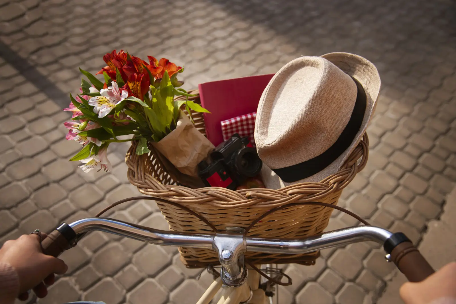 Bicycle basket filled with flowers, a straw hat, and a camera on a cobblestone street