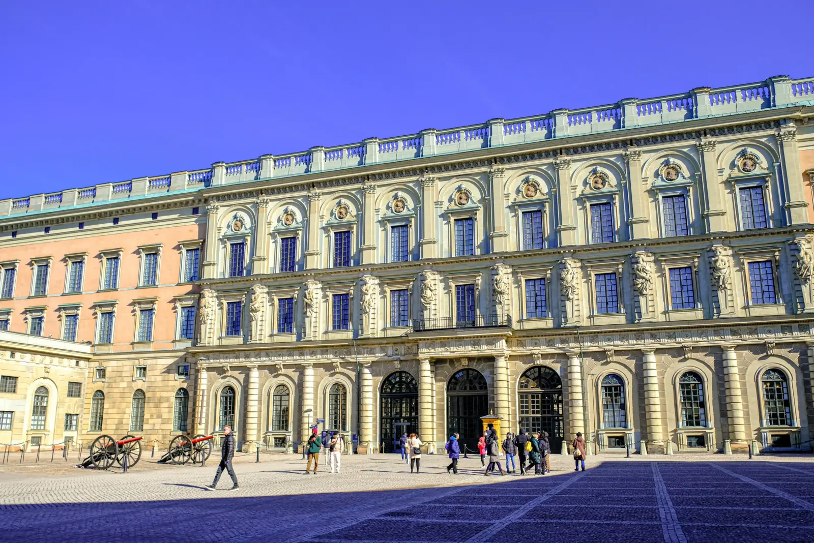 Sunlit facade of the Royal Palace courtyard with visitors and ceremonial cannons