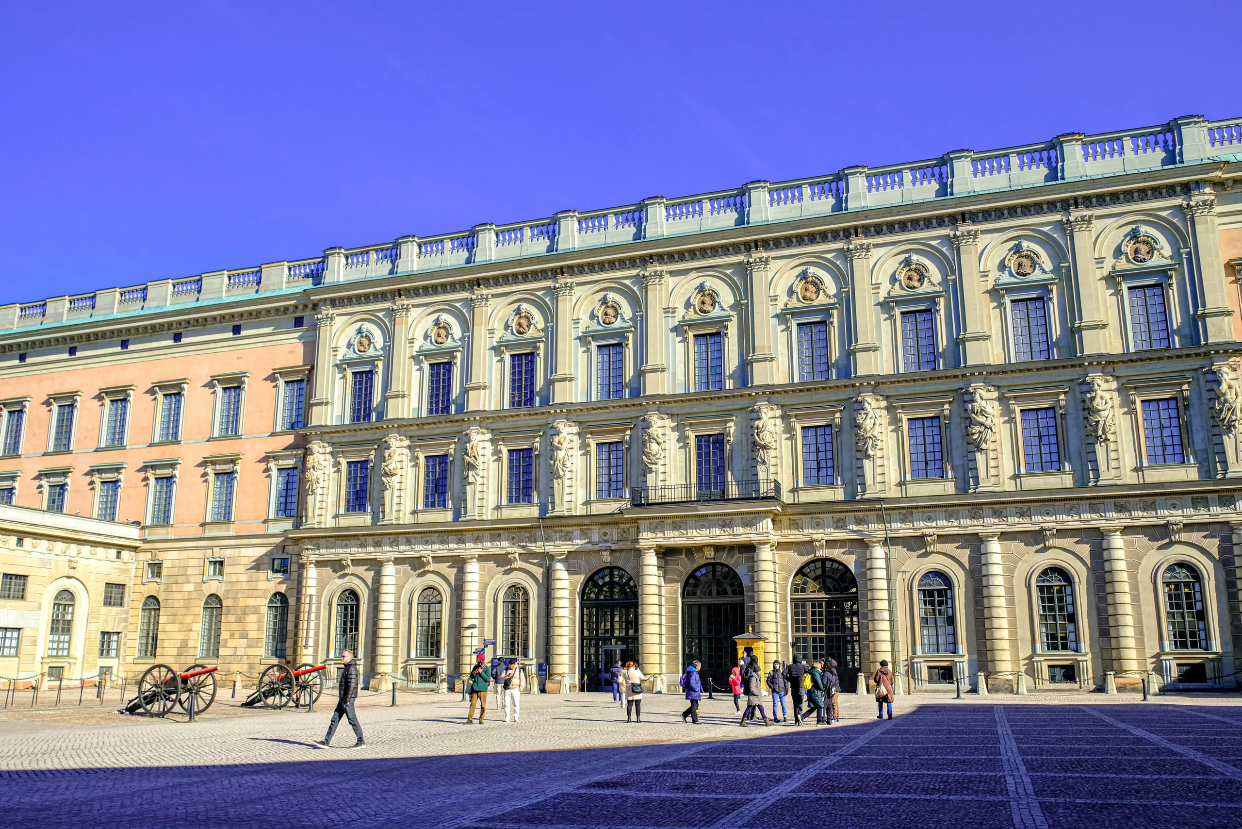 Sunlit facade of the Royal Palace courtyard with visitors and ceremonial cannons