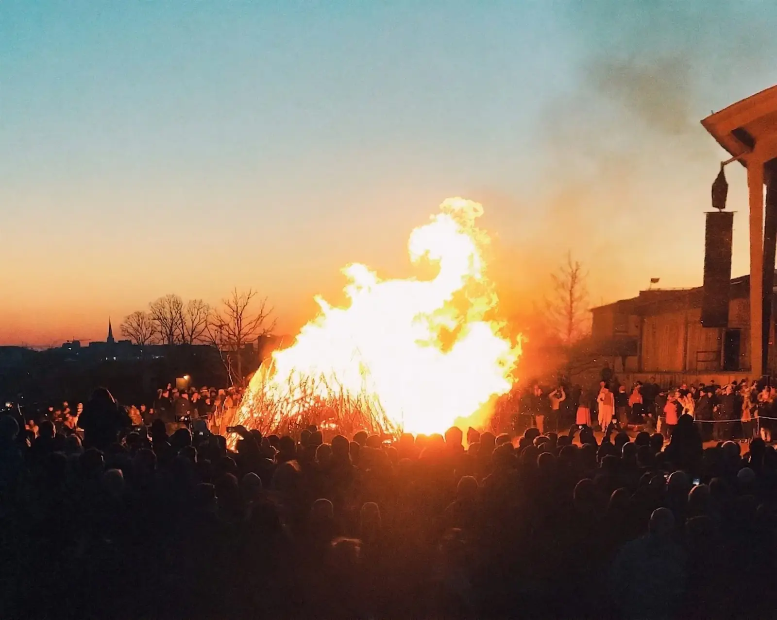 Large Valborg bonfire burning at dusk with a crowd gathered around it
